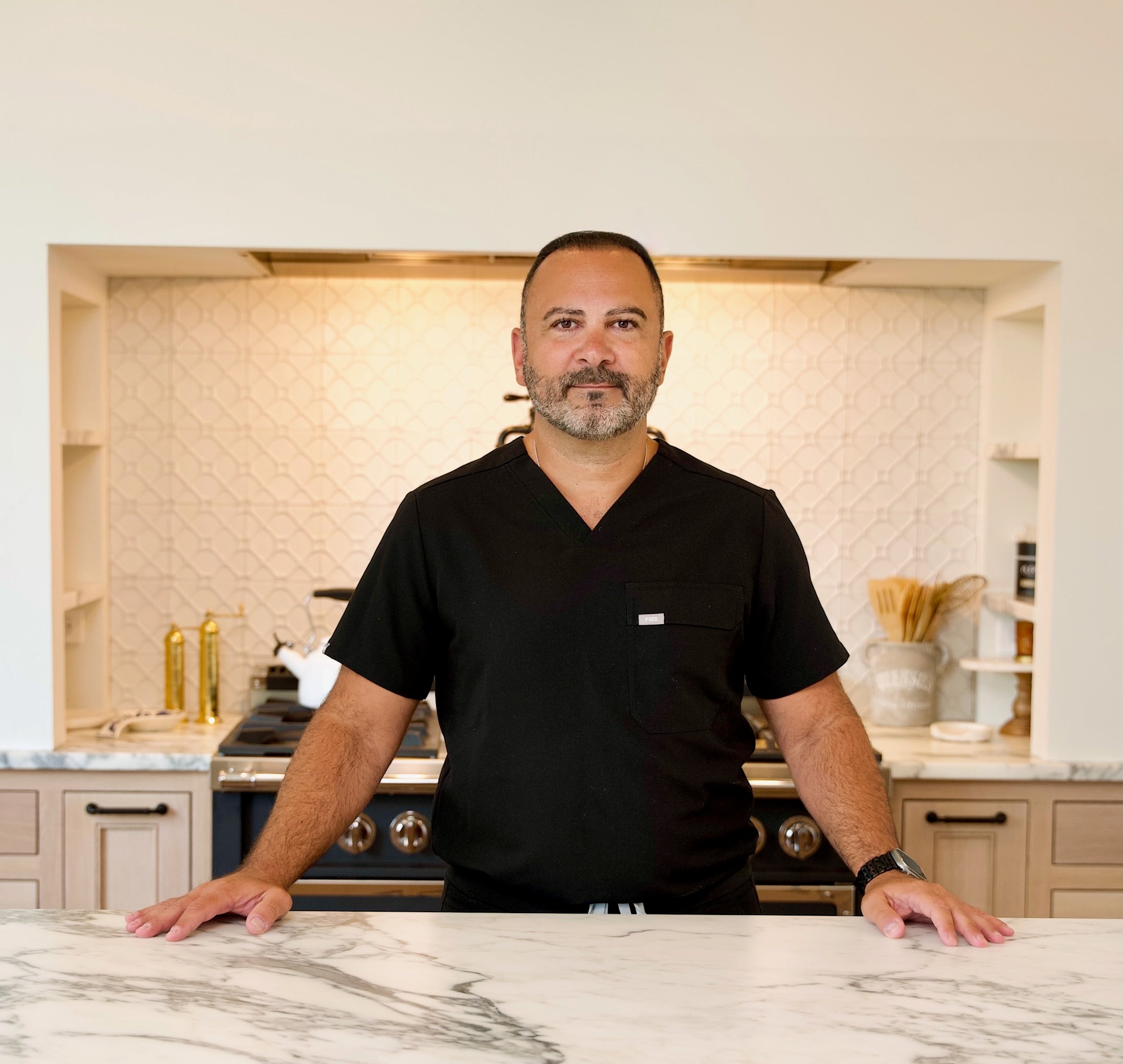 Habthera Nutrition - About Us. A doctor in a black shirt standing in a kitchen with marble countertops and tiled walls.